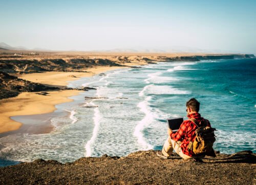 Travel and technology digital job nomad lifestyle people - man sitting on the coast work with laptop computer - wonderful beach in background - freedom from office life